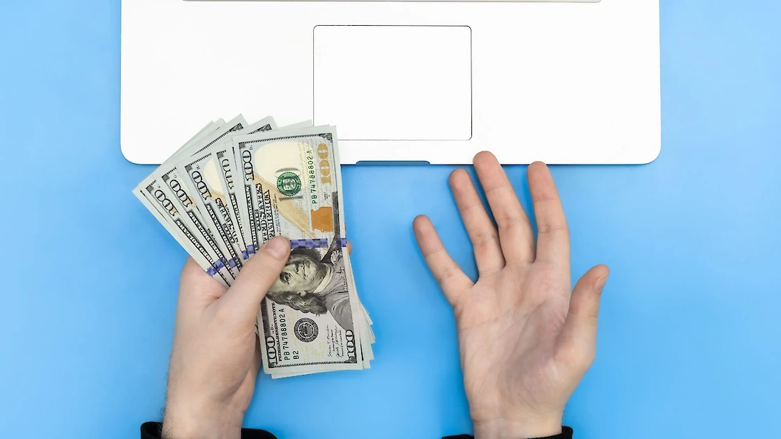 De handen van een gefrustreerd persoon die honderden dollars boven een laptop houdt.